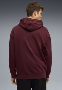 Sudadera con capucha color burdeos de corte relajado, con puños y dobladillo acanalados. Textura suave, sin patrones visibles ni detalles de herrajes.