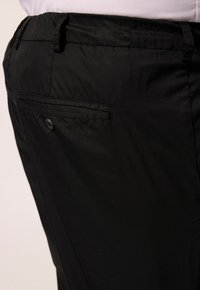 Zwarte broek met een gladde textuur, met een achterzak met knoopsluiting en riemlussen, weergegeven vanuit een zijaanzicht.