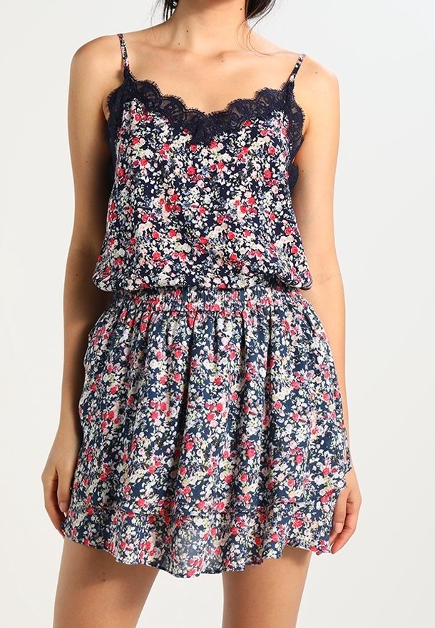 Femme portant une robe sans manches bleu marine à fleurs rouges et blanches avec une bordure en dentelle noire au niveau du décolleté.