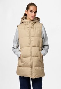 Beige Steppweste mit Kapuze, gekennzeichnet durch ein gesteppte Design, Frontreißverschluss und Seitentaschen, getragen über einem grauen Langarmshirt.