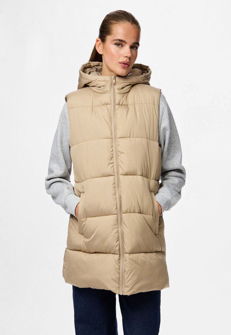 Beige Steppweste mit Kapuze, gekennzeichnet durch ein gesteppte Design, Frontreißverschluss und Seitentaschen, getragen über einem grauen Langarmshirt.