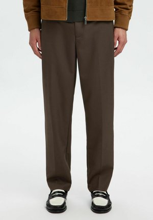 Homme portant un pantalon marron droit avec des mocassins noirs et blancs, mains reposant le long du corps, recadré à la taille et aux pieds.