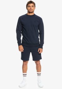 Navy blue sweatshirt with crew neck, ribbed cuffs, and hem; paired with matching shorts. White sneakers and striped socks complete the outfit.