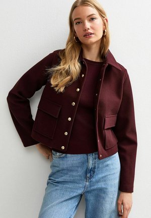 Veste courte bourgogne avec des boutons dorés et deux poches avant, associée à un haut bourgogne côtelé et un jean bleu clair. Texture lisse.
