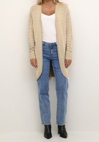 Femme portant un cardigan en maille crème par-dessus un haut blanc, un jean bleu taille haute et des bottines à talons noires, debout devant un fond uni.