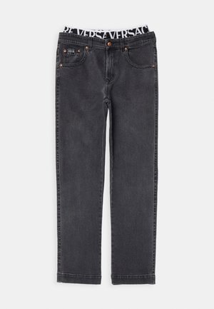 Man staat en draagt een wit T-shirt, zwarte jeans, zichtbare tailleband met de tekst "VERSA" en witte sneakers met zilveren details.