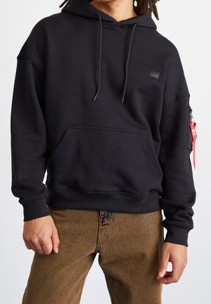 Osoba ubrana w czarną bluzę z kapturem z przednią kieszenią i troczkami, brązowe spodnie oraz czerwoną metkę przymocowaną do kieszeni na zamek na lewym ramieniu.