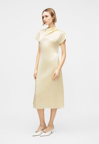 Kvinde med kort blondt hår iført en cremefarvet satin midi-kjole og hvide kitten heel-sko med ankelsnore, stående mod en ensfarvet baggrund.