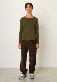 Olive green long-sleeve top with a wide neckline, paired with brown corduroy pants and gray sneakers. Smooth texture, relaxed fit.