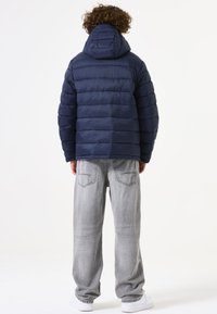 Chaqueta de plumas azul marino con capucha, diseño acolchado y puños elásticos, combinada con vaqueros baggy gris claro y zapatillas blancas.