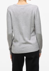 Personne portant un pull léger à manches longues gris clair et un pantalon noir, vue de dos avec les mains reposant le long du corps.