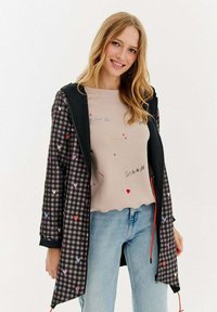 Smaidoša sieviete, kura valkā melno un rozā ķekaru jaku ar krāsainiem sirdīm, bēšas krāsas krekliņu ar maziem tekstiem un gaiši zilos džinsos, stāv pretī baltai fona.