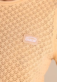 Haut tricoté orange clair avec un motif en losange, présentant une finition texturée et une étiquette brodée "Oxbow" sur le côté.