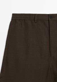 Pantalons en lin marron avec une taille élastique, une fermeture à bouton, des poches latérales et un tissu lisse et texturé.
