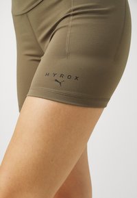 Olivengrønn tilpassede shorts laget av elastisk stoff, med svart "HYROX" logo og Puma-emblem ved den nederste kanten, samt synlige sømdetaljer.