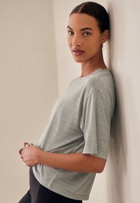 Heleroheline lühikeste varrukatega t-särk, millel on peen tekstuurmuster, lõdvestunud lõige ja ümar kaelus, kantud mustade leggingsidega seina vastu.