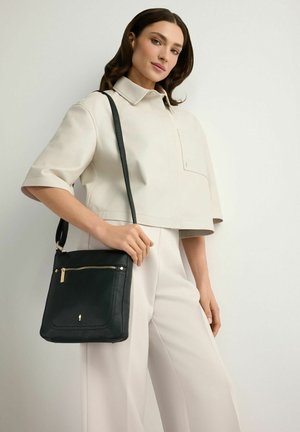 Kobieta w beżowym stroju pozuje, trzymając czarną skórzaną torebkę na ramię z przednią kieszenią na suwak na jednolitym tle.