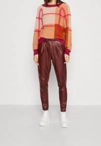 Pull en tricot texturé et coloré avec des motifs géométriques dans des tons pêche et rose ; porté avec un jogging en cuir bordeaux foncé et des baskets blanches.