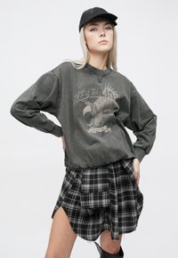 Felpa grigia oversize con grafica di un'aquila, abbinata a una gonna a quadri nera e grigia con tasca e orlo sfrangiato. Cappellino nero indossato.