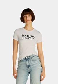 T-shirt bianco a maniche corte con "tommy jeans" stampato in corsivo blu navy. Abbinato a jeans a vita alta blu chiaro.