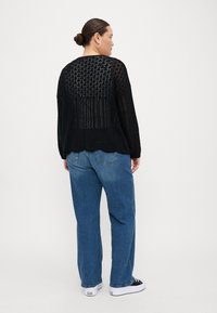 Black open-knit sweater with a loose fit, scalloped hem, and back detail. Paired with light blue jeans and black sneakers.