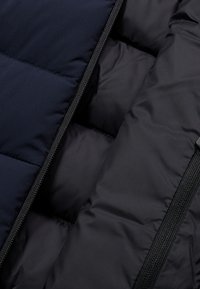 Schwarze Daunenjacke mit navyfarbenen Abschnitten, gestepptem Stoff und sichtbarem Reißverschluss. Das Außenmaterial ist glatt und wasserabweisend.