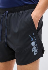 Άνδρας που φοράει μαύρο σορτς τρεξίματος Nike, με το χέρι στην τσέπη, λογότυπο Nike και το κείμενο "Nike" ορατά στον αριστερό μηρό και τη μέση.