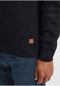 Maglione nero a maglia con un motivo a costine testurizzato. Presenta una toppa in pelle marrone con logo sull'orlo e polsini aderenti. Indossato con jeans blu.