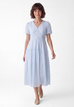 Frau mit kurzen braunen Haaren, die ein hellblaues Midi-Kleid mit Puffärmeln und beigen flachen Schuhen trägt, geht vorwärts auf weißem Hintergrund.