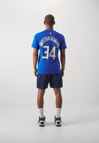 Modrá basketbalová dres s menom "ANTETOKOUNMPO" a číslom "34" vytlačeným zlatou farbou na chrbte, v kombinácii s námorníckymi šortkami a čiernymi topánkami.