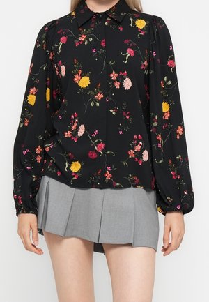 Sort bluse med lange ærmer og flerfarvet blomsterprint kombineret med en kort grå plisseret nederdel, båret af en person med lys hud og glat blondt hår.