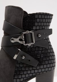 Acercamiento de una bota negra hasta el tobillo con un patrón cuadrado texturizado, dos correas de piel y detalles de hebilla plateados.