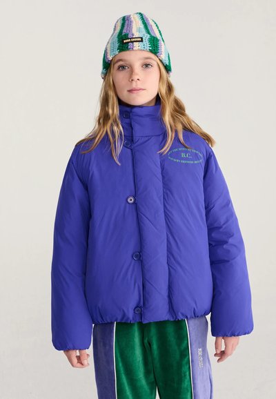 Fille aux longs cheveux porte une doudoune bleue, un pantalon en velours vert et bleu et un bonnet tricoté rayé avec une étiquette noire.