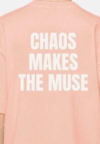 Camiseta de algodón rosa con texto grande en blanco en la espalda que dice "EL CAOS INSPIRA A LA MUSA." Mangas cortas y corte relajado.