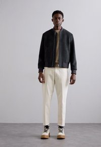 Chaqueta bomber verde oscura, suéter beige, pantalones crema y botas beige con cordones negros. Textura suave, silueta ajustada, diseño minimalista.
