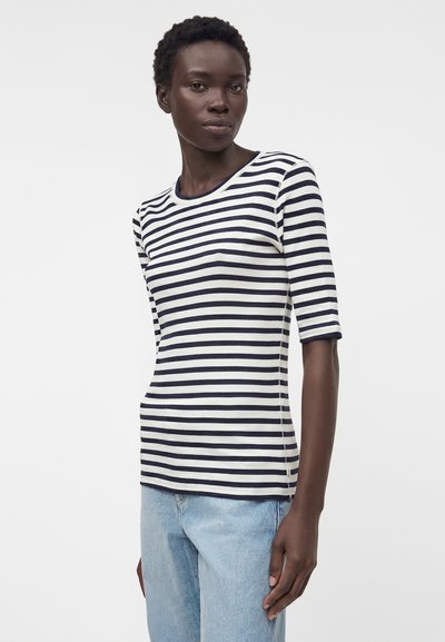 Γυναίκα που φοράει εφαρμοστή μαύρο-λευκή ριγέ μπλούζα με μανίκια ως το αγκώνα και ανοιχτόχρωμα μπλε τζιν, στέκεται σε απλό λευκό φόντο.