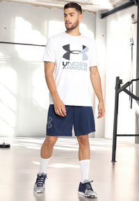 White short-sleeve T-shirt with dark gray Under Armour logo, paired with navy blue shorts featuring a logo pattern; white socks and athletic shoes.