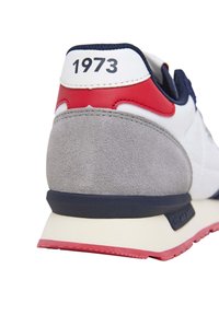 Vit sneaker med grå mockaaccenter, röda och marinblå detaljer. Har en platt gummisula, texturerad yta och "1973" tryckt på hälen.