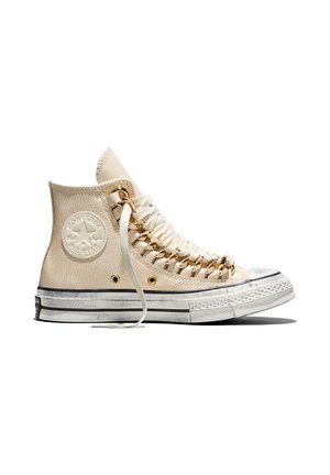 Zapatilla alta Converse beige con suela blanca gruesa, ojales de metal dorado, cordones anchos color crema y logo Converse en relieve en el lateral.
