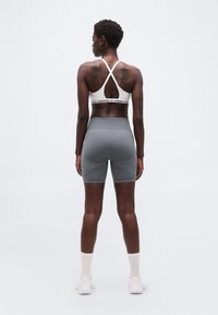 Γυναίκα που στέκεται με την πλάτη στραμμένη, φοράει λευκό αθλητικό σουτιέν με διασταυρωμένους ιμάντες, γκρι εφαρμοστά σορτς, λευκές κάλτσες και λευκά αθλητικά παπούτσια.