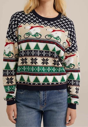 Femme portant un pull festif avec des ours polaires chevauchant des vélos verts, des sapins de Noël et des motifs de flocons de neige en rouge, vert, marine et beige.