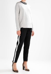 Persoon met een lichtgrijs topje met lange mouwen, zwarte broek met witte zijstrepen, en hoge hakken met luipaardprint, staande op een witte achtergrond.