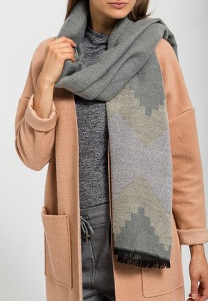 Femme portant un manteau beige, un pull gris et une écharpe grise à motifs enroulée autour du cou, tenant l'écharpe d'une main.