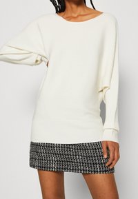 Witte geribbelde trui met brede bootnek en omlaag gevallen schouders, gecombineerd met een zwart-wit gestructureerde minirok. Zachte gebreide stof.