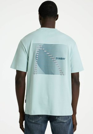 Man draagt een lichtblauw T-shirt met een lijnvormige afbeelding en het woord "CHASIN'" op de achterkant, gecombineerd met een blauwe spijkerbroek.