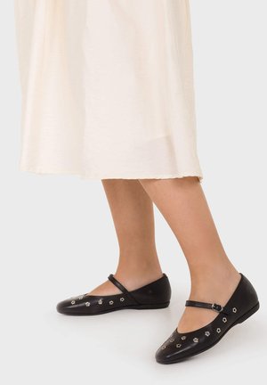 Nogi w kremowej spódnicy midi i czarnych balerinach Mary Jane z małymi srebrnymi kwiatowymi ćwiekami na białym tle.