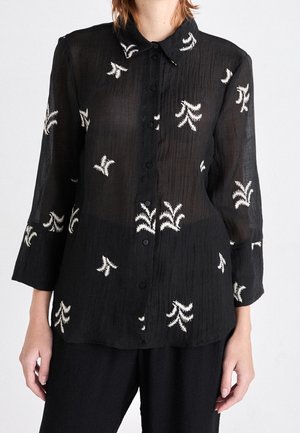 Blouse noire boutonnée en tissu texturé avec des motifs floraux abstraits brodés en blanc, portée avec un pantalon noir assorti.