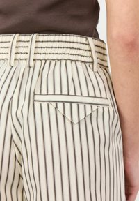Striped high-waisted trousers in cream and brown fabric, featuring a ribbed elastic waistband and a distinctive angled back pocket detail.