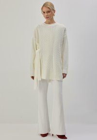 Pull en tricot blanc avec motifs torsadés texturés, doté de liens sur les côtés. Associé à un pantalon blanc évasé et des chaussures à talons rouges. Fond uni.