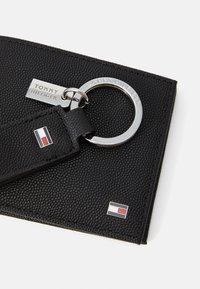 Porte-clés en cuir noir texturé avec un anneau en métal argenté, gravé "TOMMY HILFIGER", avec un patch de logo rouge, blanc et bleu.